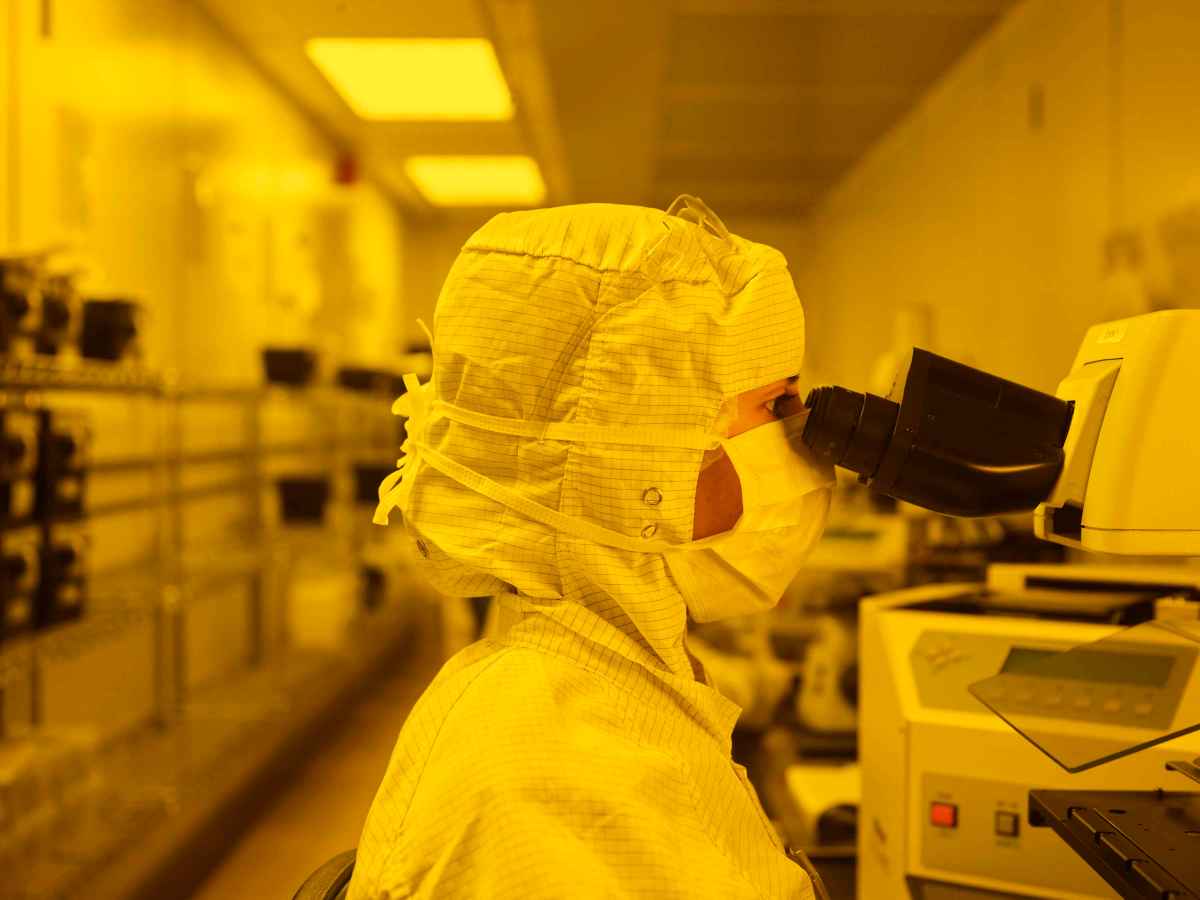 Woman wearing PPE in a clean room with a yellow tint, looking into microscope.